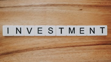 The word INVESTMENT spelled out in black letters on white Scrabble-style tiles placed on a wooden surface.