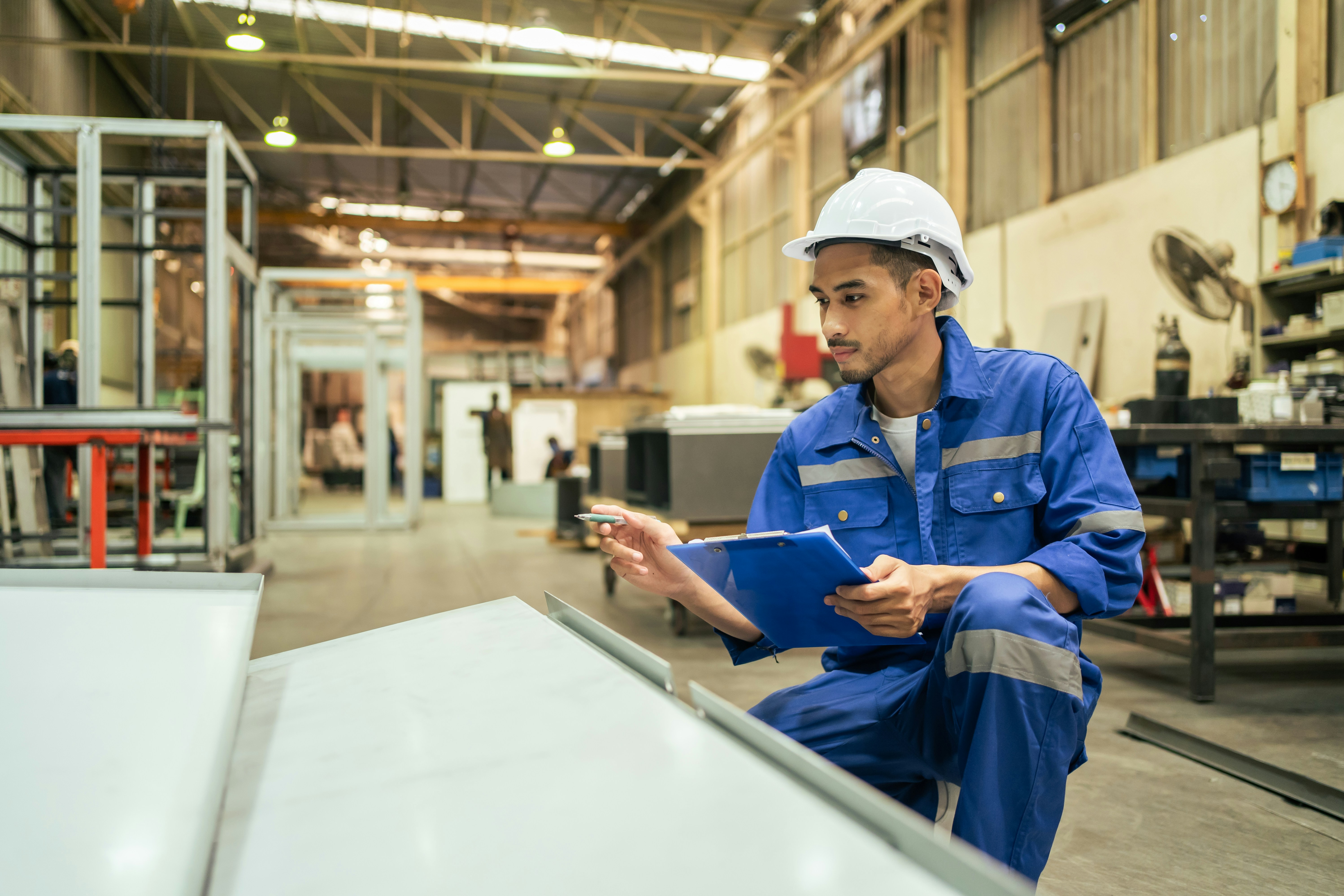 Man kneeling in warehouse with a clipboard calculating something, wearing hardhad, holding a clipboard and a pencil. 